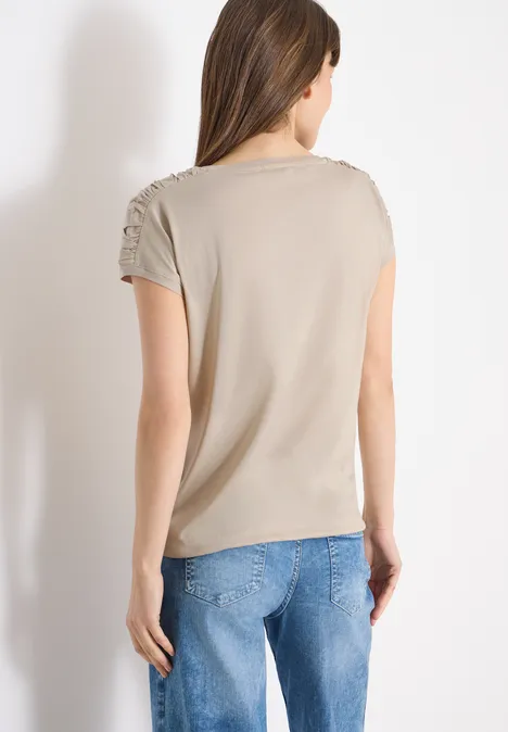 Frau von hinten in beiger, kurzärmeliger Bluse mit Raffungen und blauen Jeans vor weißer Wand.